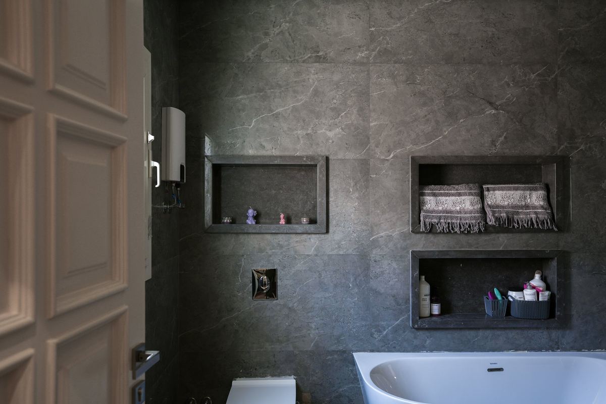 After: L-Villa finished bathroom: grey stone-look tiles, lit niches with towels and a freestanding tub